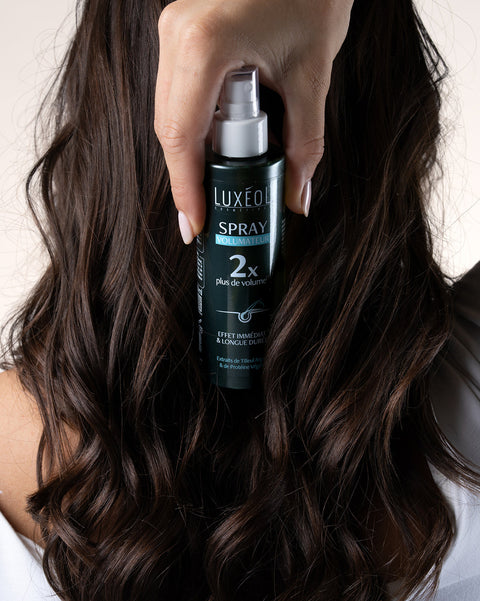 Person holding a Luxeux hair spray bottle with long, wavy hair.